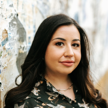 Woman with long dark hair standing beside textured wall, looking at camera.